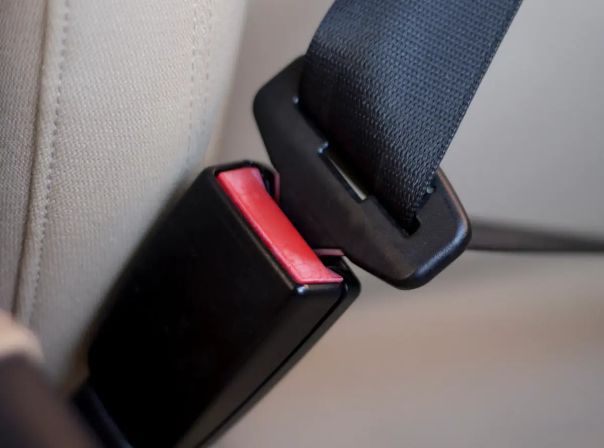 Black seat belt buckle with red release beside a latch tongue held in a pillar loop on a beige seat