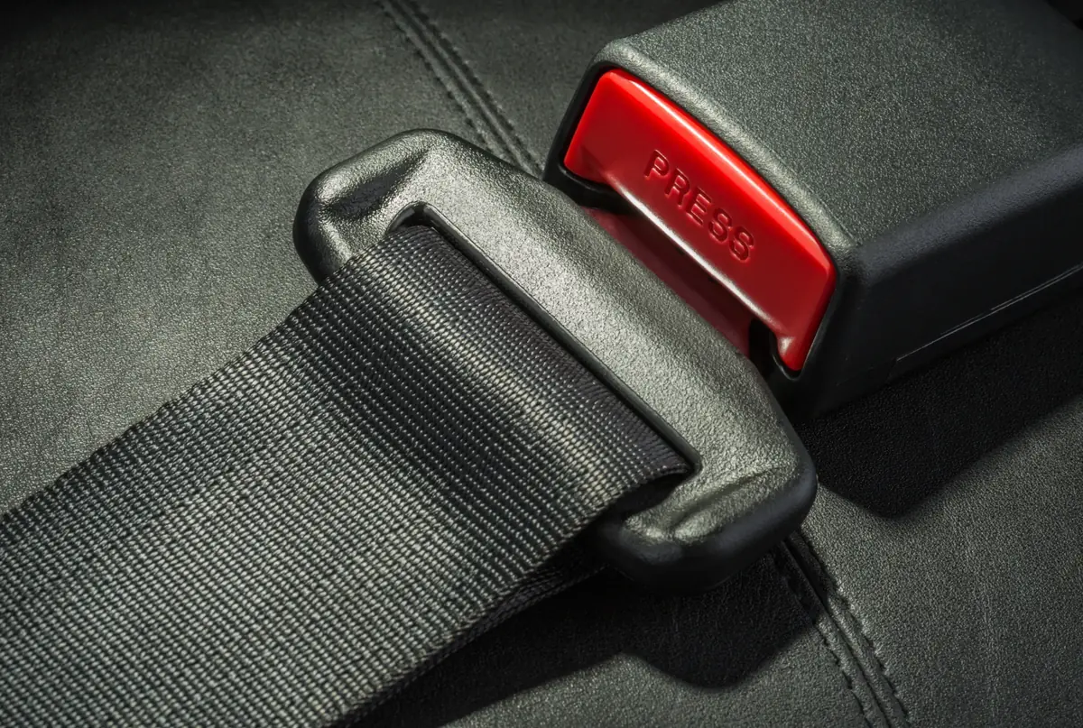 Close-up of a black seat belt webbing, metal latch tongue and buckle with a red press release, engaged on a vehicle seat