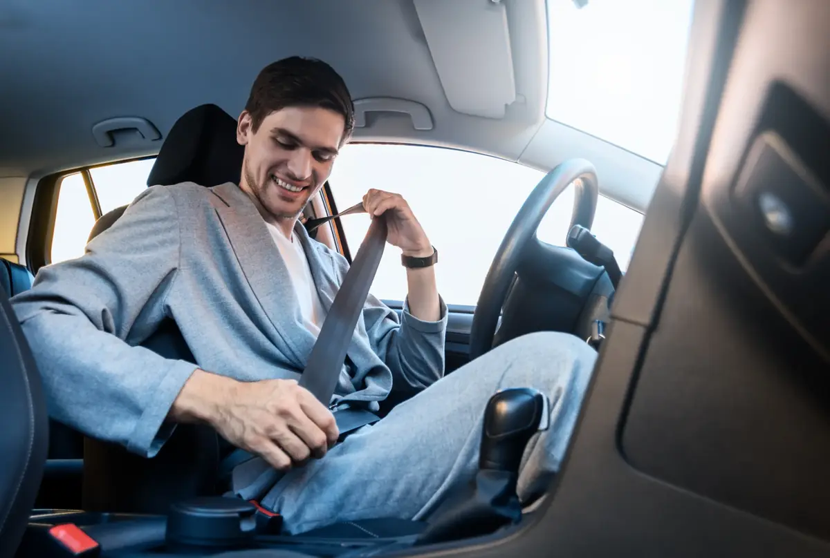 Occupant in a grey jacket drawing a seat belt across the body from the pillar inside a car