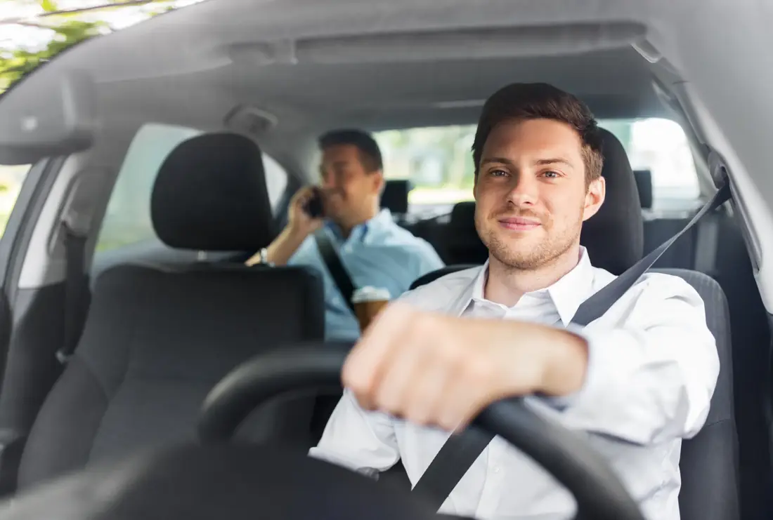 Belted driver at the wheel of a vehicle with a belted passenger seated behind holding a phone